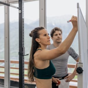 Female fitness coach writes on whiteboard as a male athlete watches in a gym.