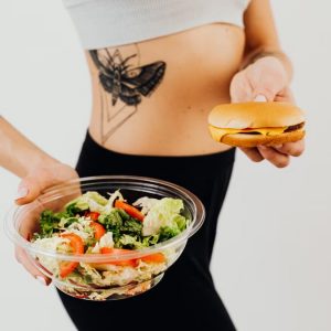 A woman with a tattoo holding a healthy vegetable salad and a cheeseburger, representing food choices.