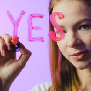 Teenage girl writing the word 'Yes' on glass with marker in studio setting.